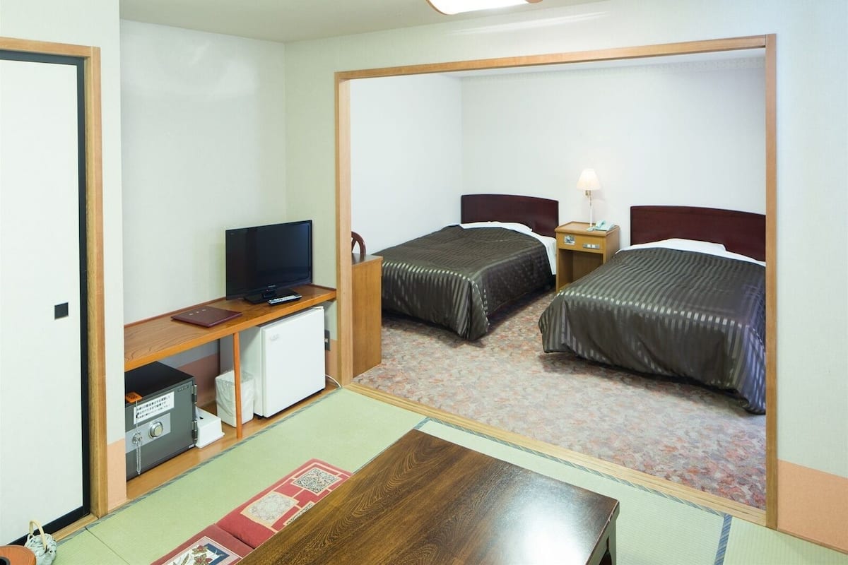 Habitación de hotel con dos camas, un escritorio con televisor, un refrigerador pequeño y una mesita de noche con lámpara.