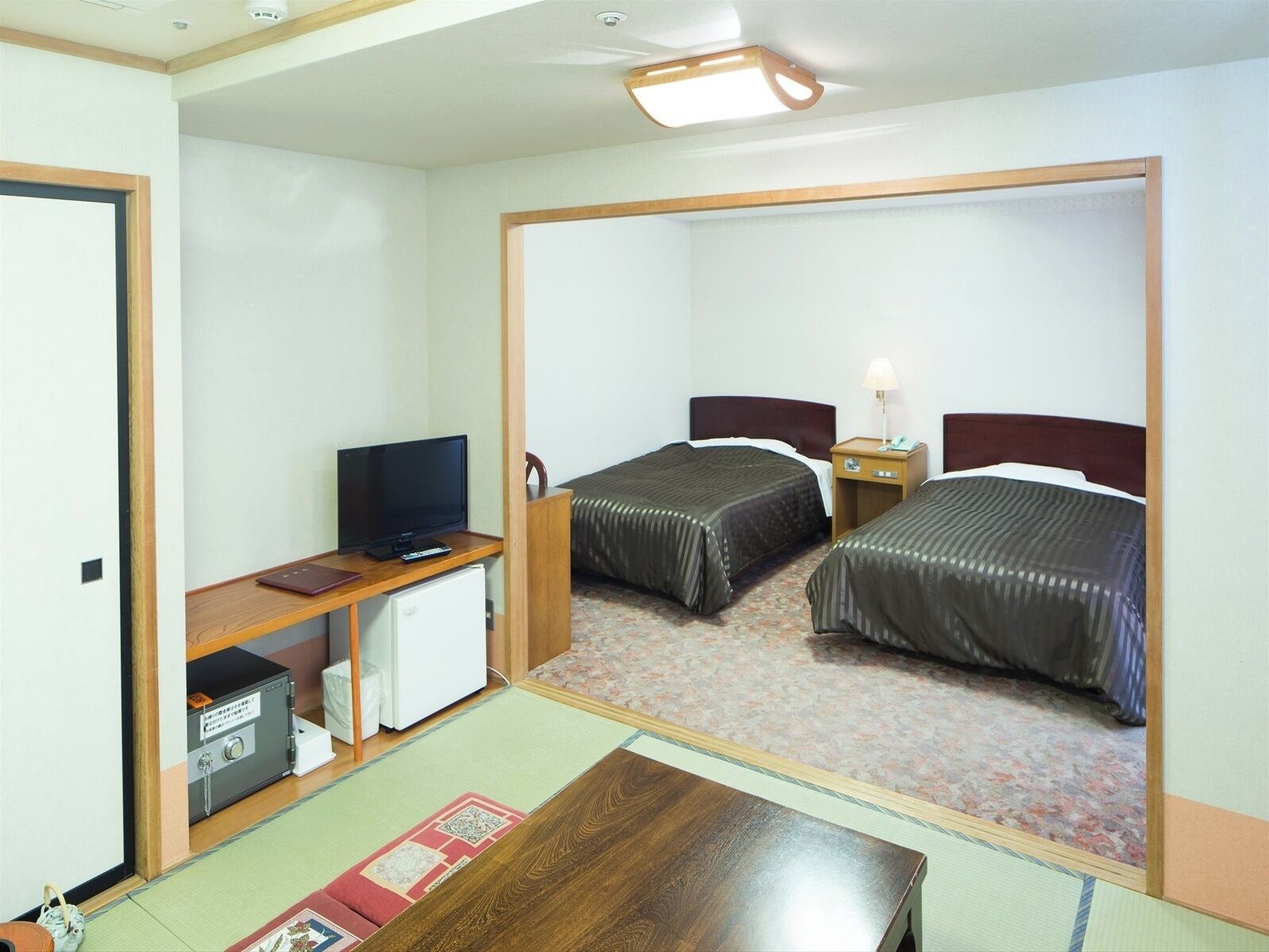 Quarto de hotel com duas camas, uma mesa de trabalho com televisão, um frigorífico pequeno e uma mesa de cabeceira com candeeiro.