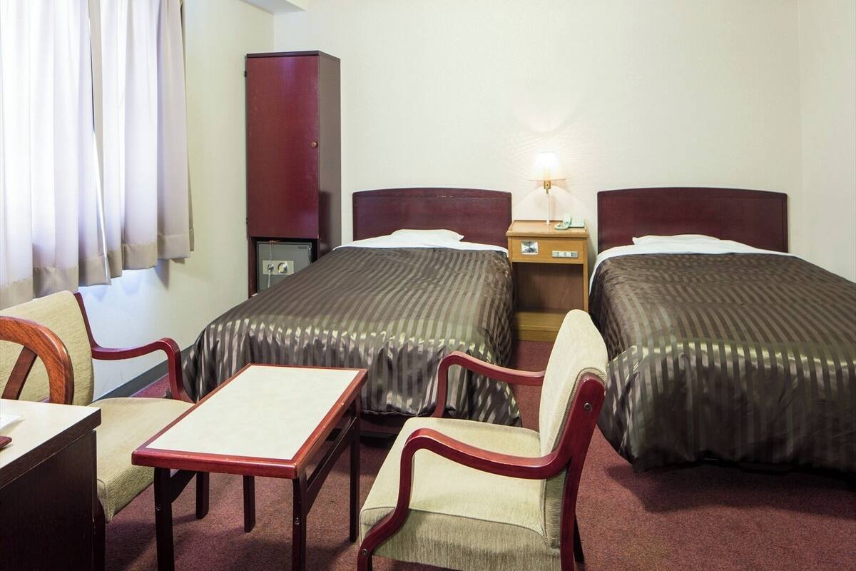 Una habitación de hotel con dos camas, un escritorio, una silla, un mueble pequeño y una mesita de noche con lámpara.