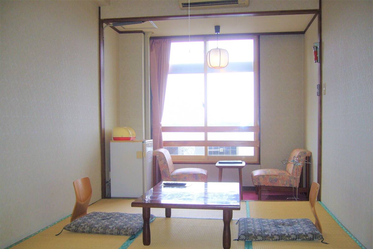 A traditional Japanese room with tatami flooring, a low table, two chairs, a small table, and a window with curtains.