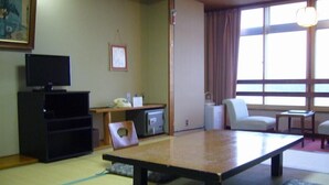 A traditional tatami mat room with a tatami mat table, a television, a small cabinet, a chair, and a window with curtains. - Sado Heights Hotel Aoki (Sado)