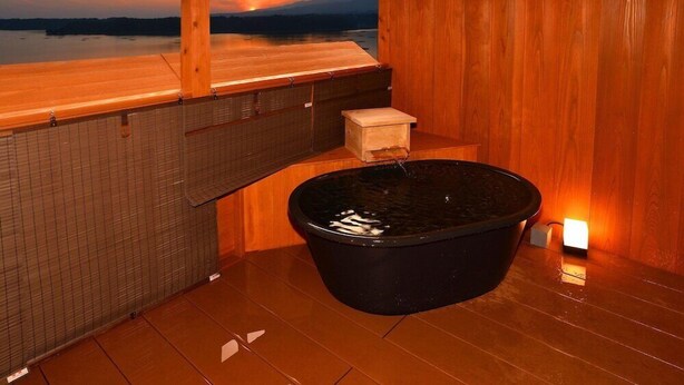 A wooden sauna with a large black tub, a small wooden box, and a view of a sunset over water. - Sado Heights Hotel Aoki (Sado)