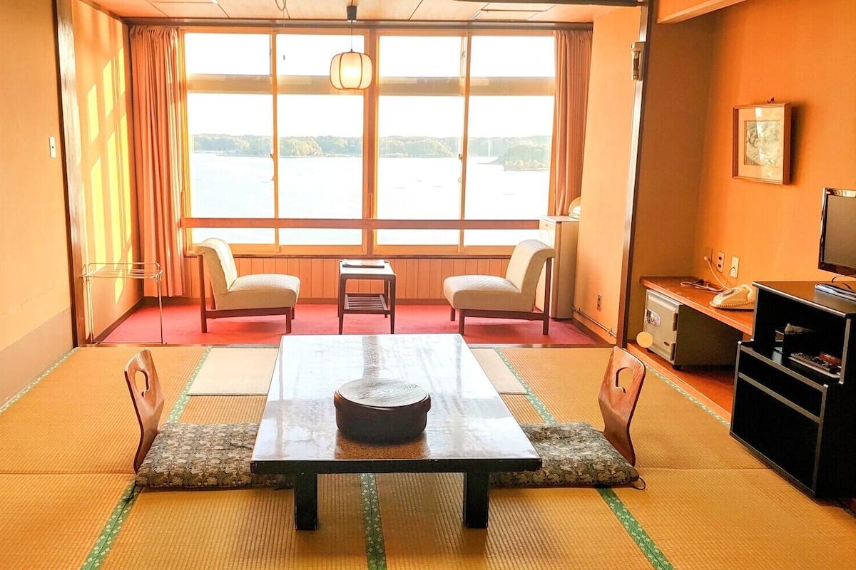 A traditional Japanese room with tatami flooring, a low table with a bowl, two chairs, a small table, a television, and a view of a body of water through large windows.