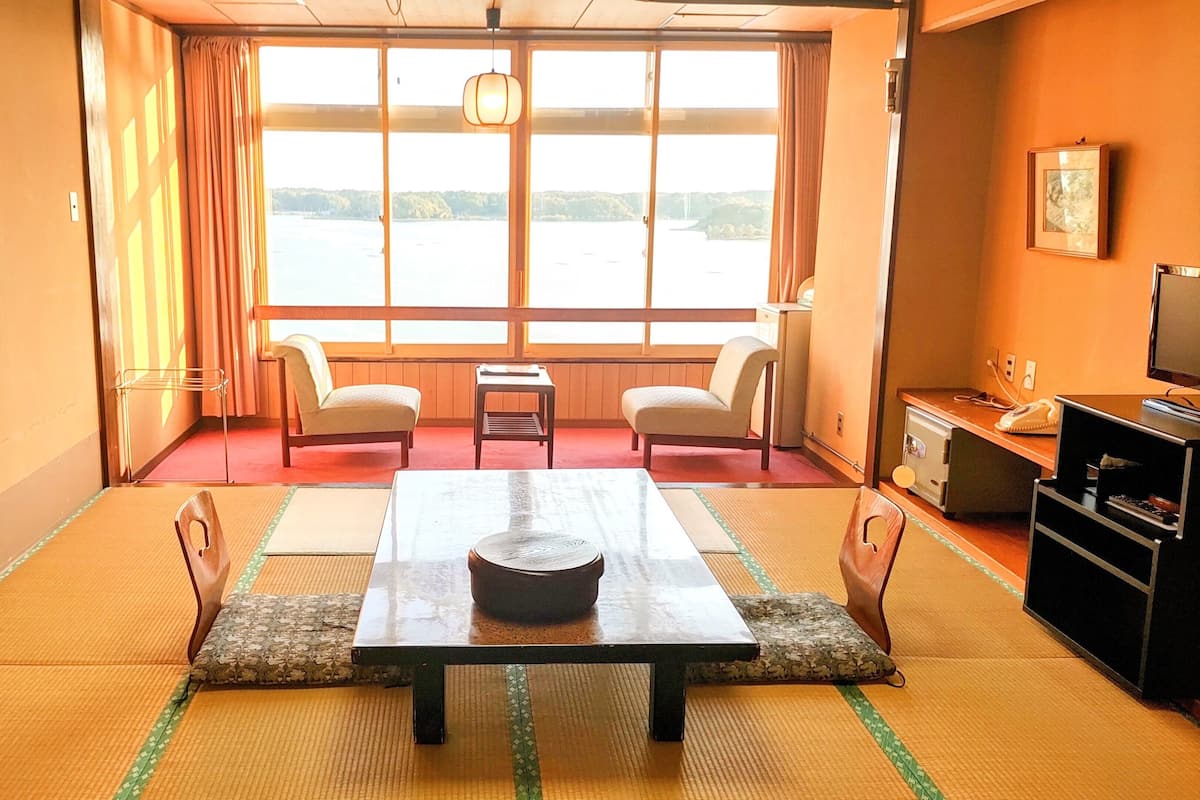 A room with tatami mats, a low table, two chairs, a television, and a view of a body of water.