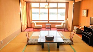 A traditional Japanese-style room with tatami flooring, a low table with a bowl, two chairs, a small table, a television, and a view of a lake through large windows. - Sado Heights Hotel Aoki (Sado)