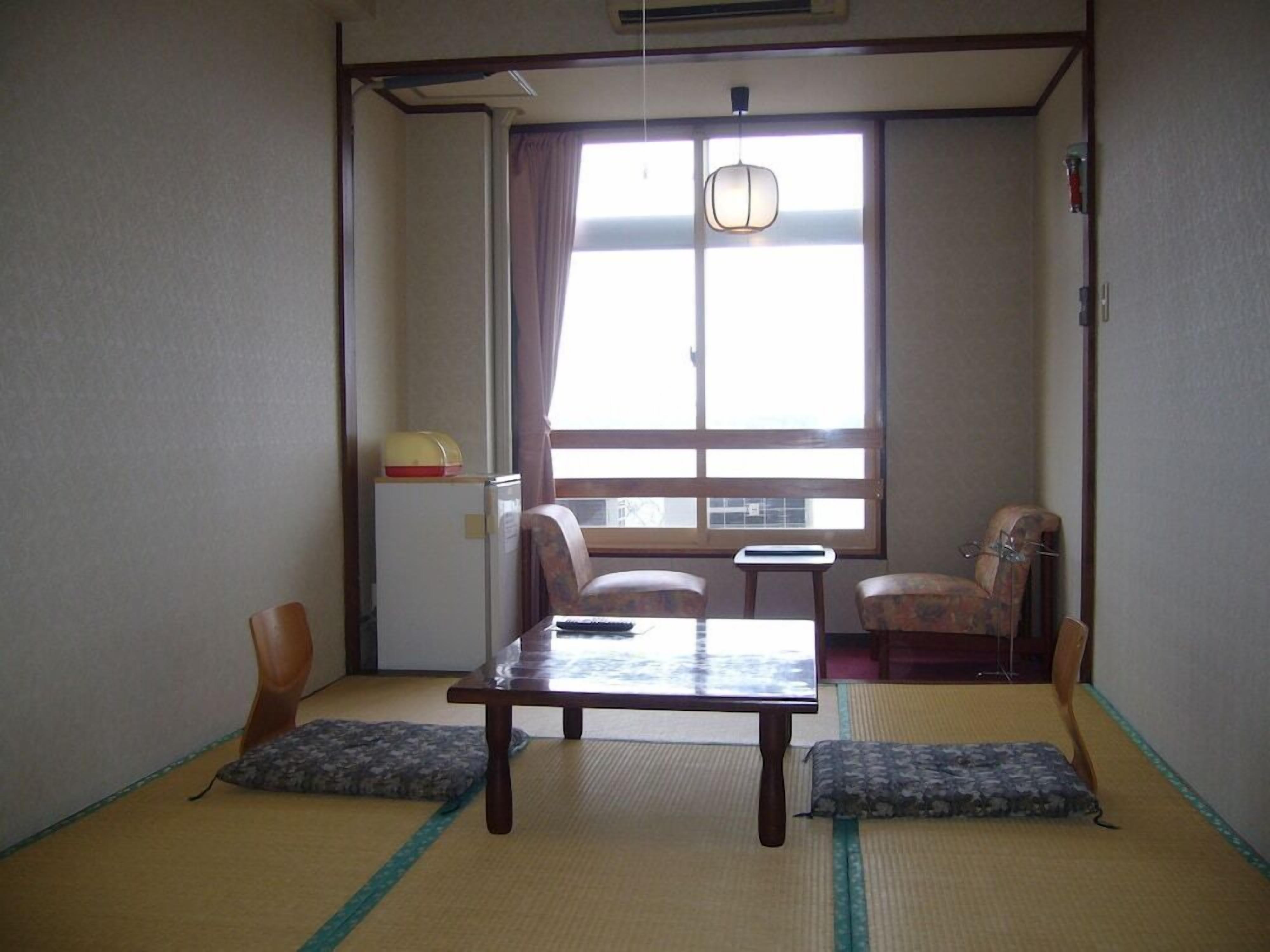 A traditional Japanese room with tatami flooring, a low table, two chairs, a small table, and a window with a view.