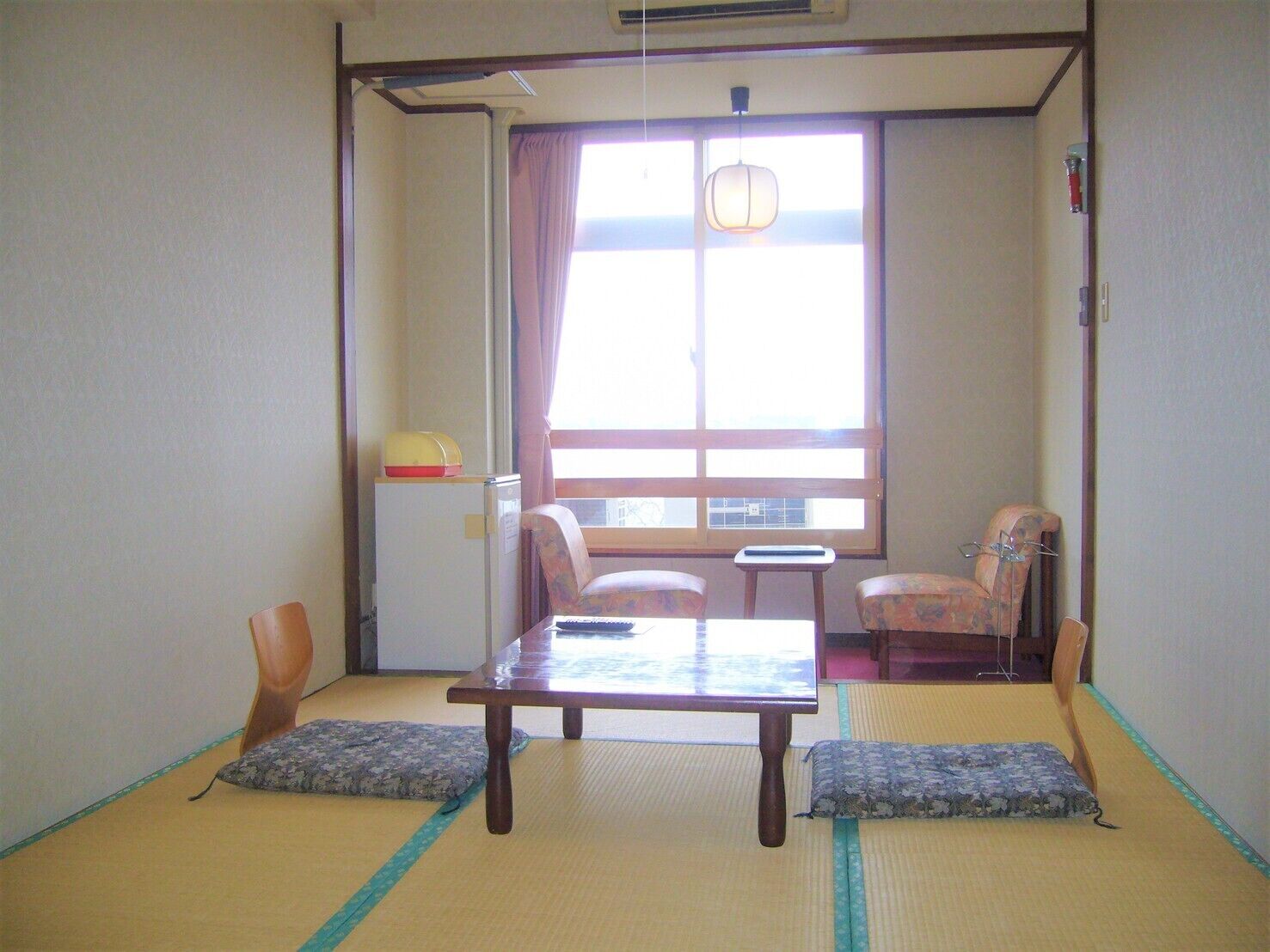 Habitació japonesa tradicional amb fons de tatami, una taula baixa, dues cadires, una taula petita i una finestra amb cortines.
