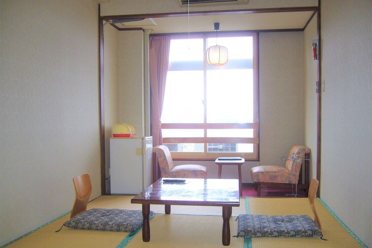 A traditional Japanese room with tatami flooring, a low table, two chairs, a small table, and a window with curtains.