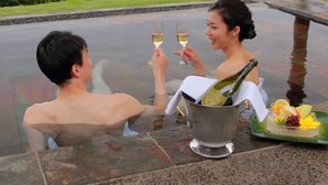 Two people toasting with champagne glasses at a table overlooking a body of water, with a fruit platter and a bottle of champagne on the table. - Seaside Resort Tsukino Usagi (Ito)
