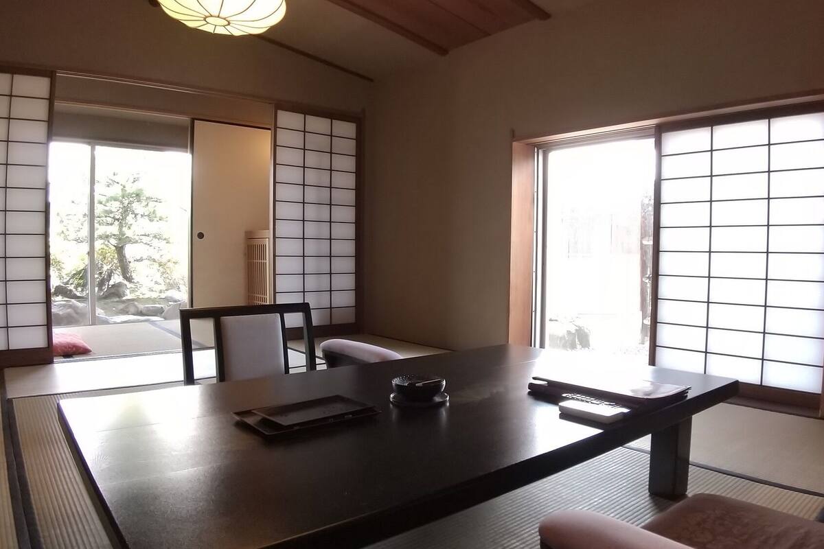 Um quarto japonês tradicional com tatames, uma mesa baixa, uma cadeira e portas deslizantes com vista para um jardim.