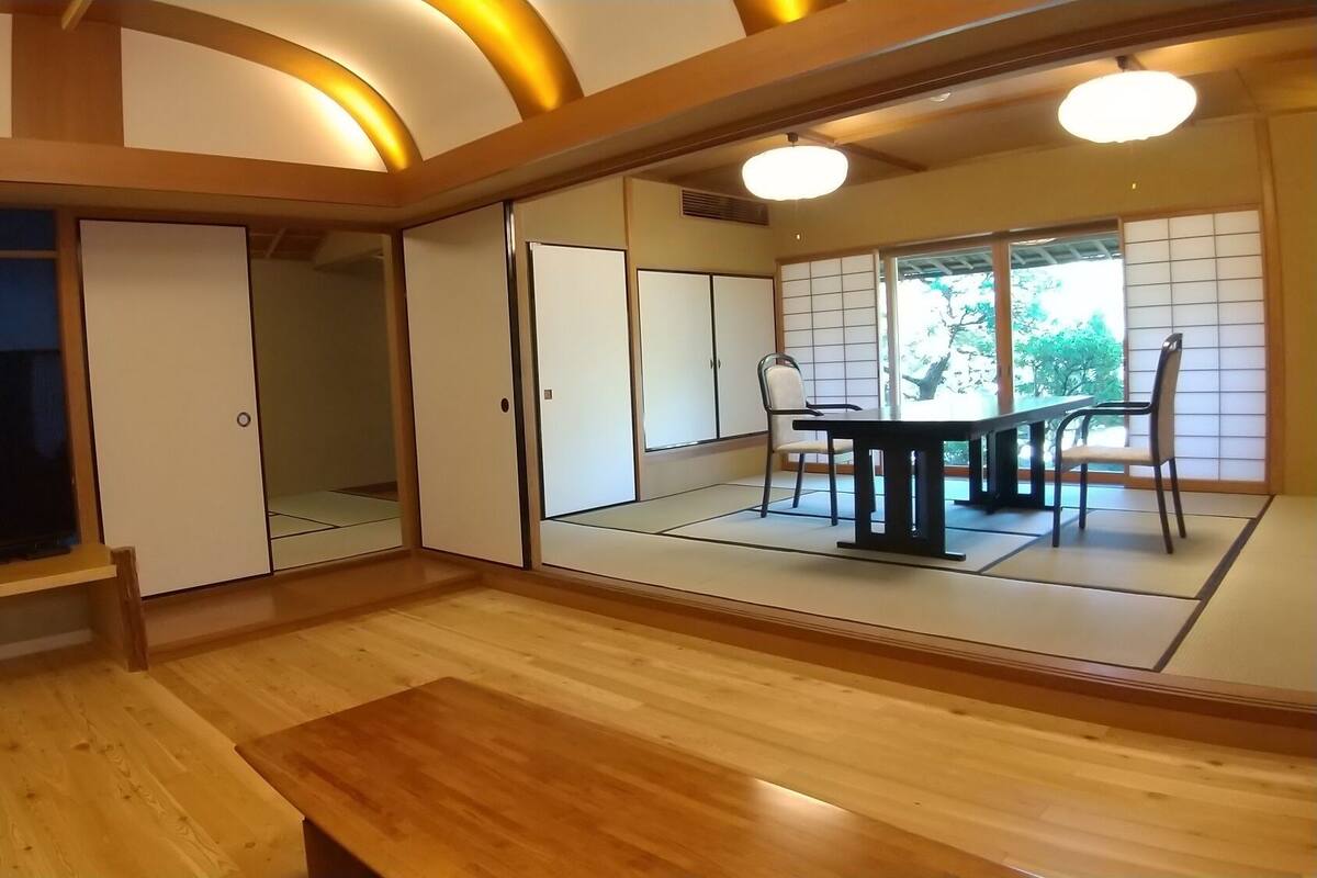 A room with a TV, sliding doors, a dining table with chairs, and a view of greenery outside.