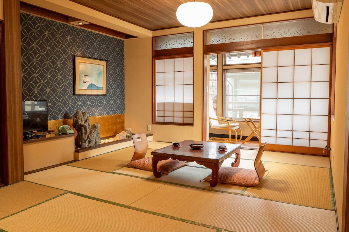 Une pièce de style japonais traditionnel, avec un sol recouvert de tatamis, une table basse avec des coussins, un téléviseur et un tableau accroché au mur.