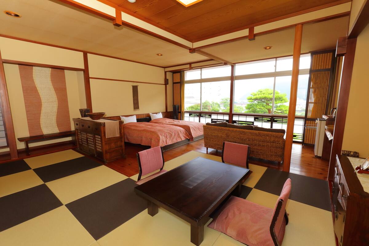 Una habitación de estilo japonés tradicional con suelo de tatami, una cama con ropa de cama rosa, una mesa de madera y vistas al exterior a través de amplias ventanas.