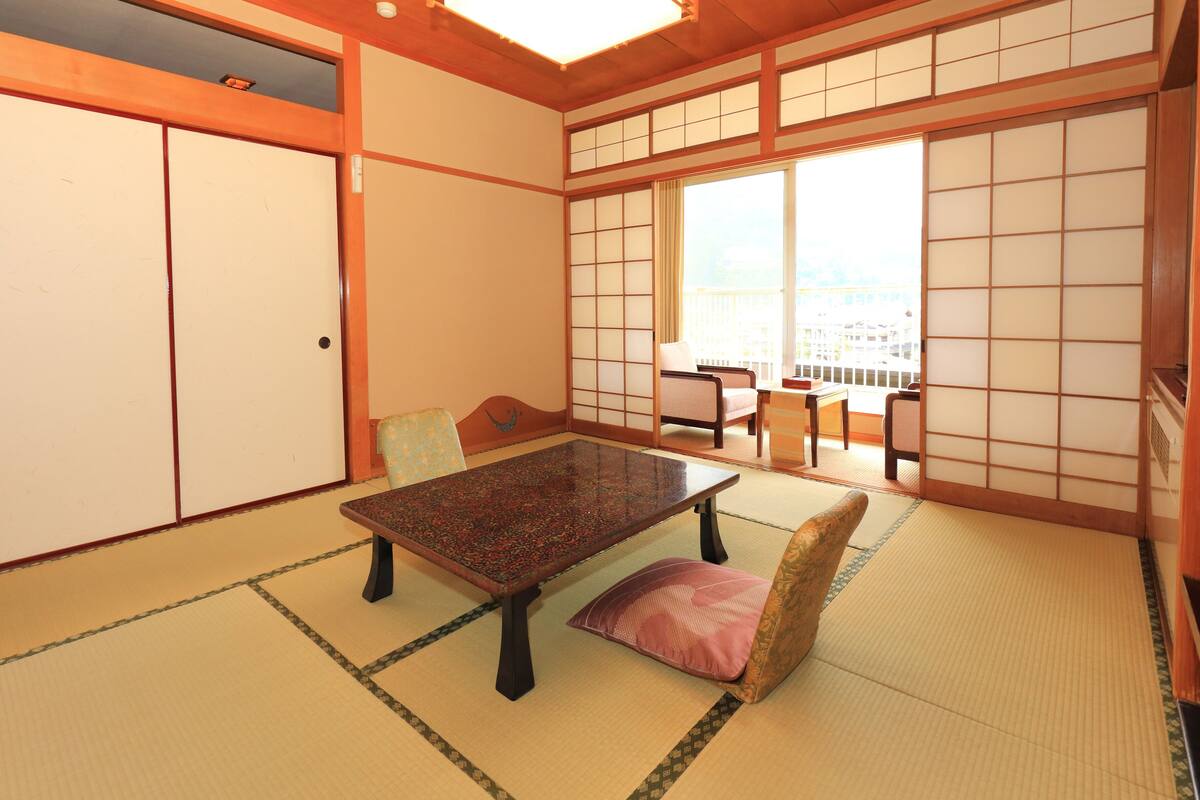 Ein traditioneller japanischer Raum mit Tatami-Böden, einem niedrigen Tisch, einer gepolsterten Sitzbank, Schiebetüren und Blick auf eine Stadtlandschaft durch große Fenster.