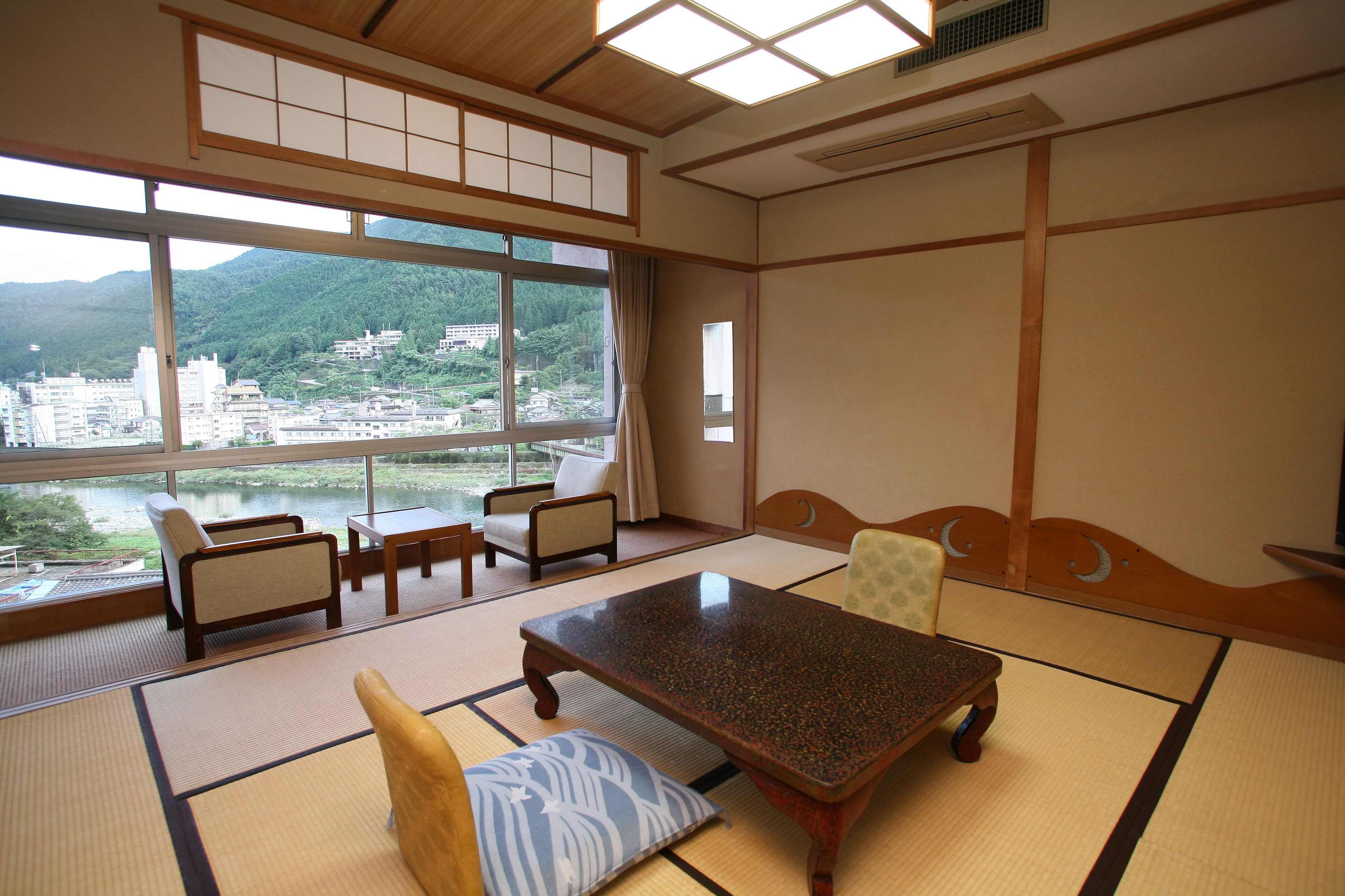 A traditional Japanese room with tatami flooring, a low wooden table, and chairs. A large window offers a view of a city and mountains.