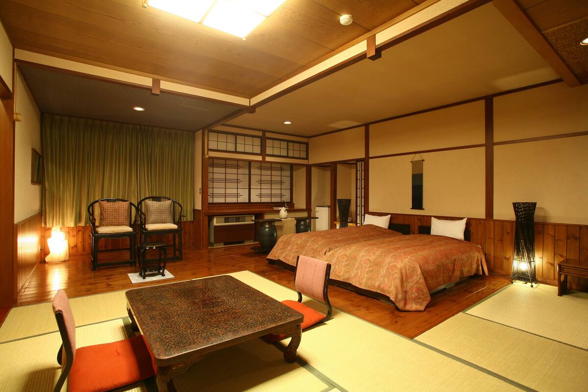 Ein traditioneller japanischer Raum mit Tatami-Boden, einem niedrigen Tisch, zwei Stühlen, einem Bett und einem Fenster mit Shoji-Bildschirmen.