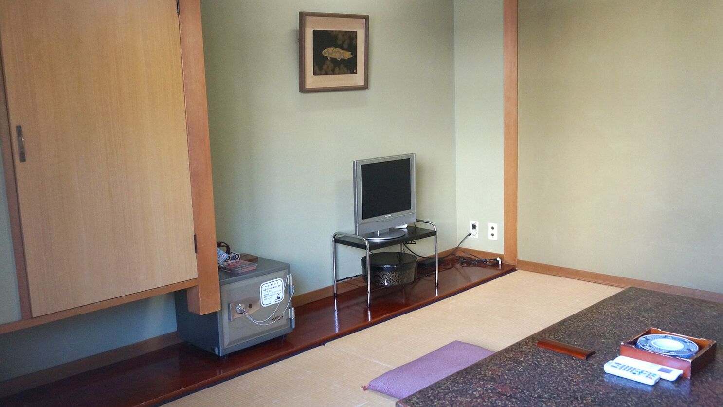 Een kleine kamer met een houten kast, een televisie op een standaard, een klein tafeltje met een afstandsbediening en een lijst aan de muur.