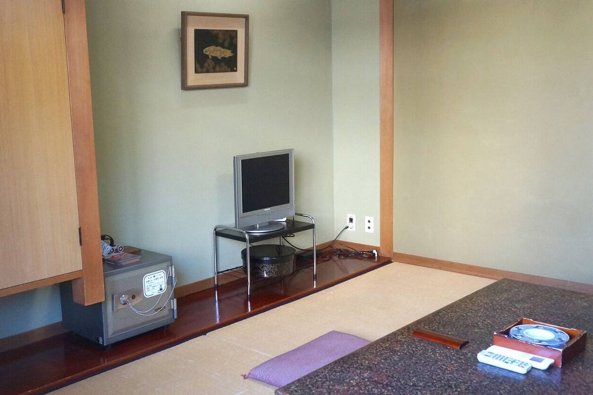 Ein kleines Zimmer mit einem Holzkasten, einem Fernseher auf einem Standfuß, einem kleinen Tisch mit einer Fernbedienung und einem gerahmten Bild an der Wand.