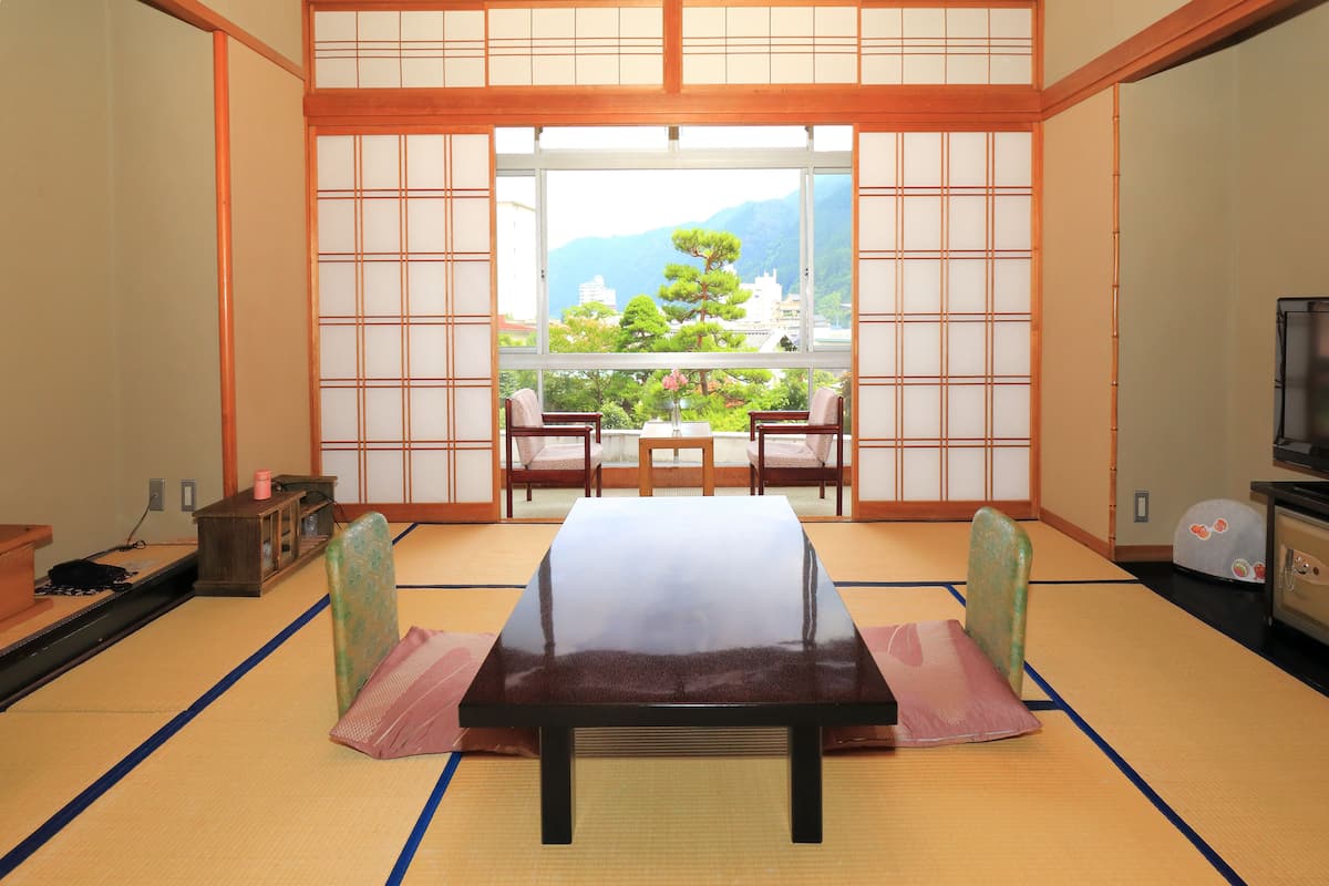 Una habitación japonesa tradicional con suelo de tatami, una mesa baja y vistas a un jardín y edificios a través de un shoji.