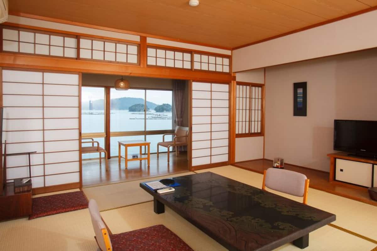 A traditional Japanese room with tatami flooring, a low table, chairs, a TV, and a view of water and mountains.