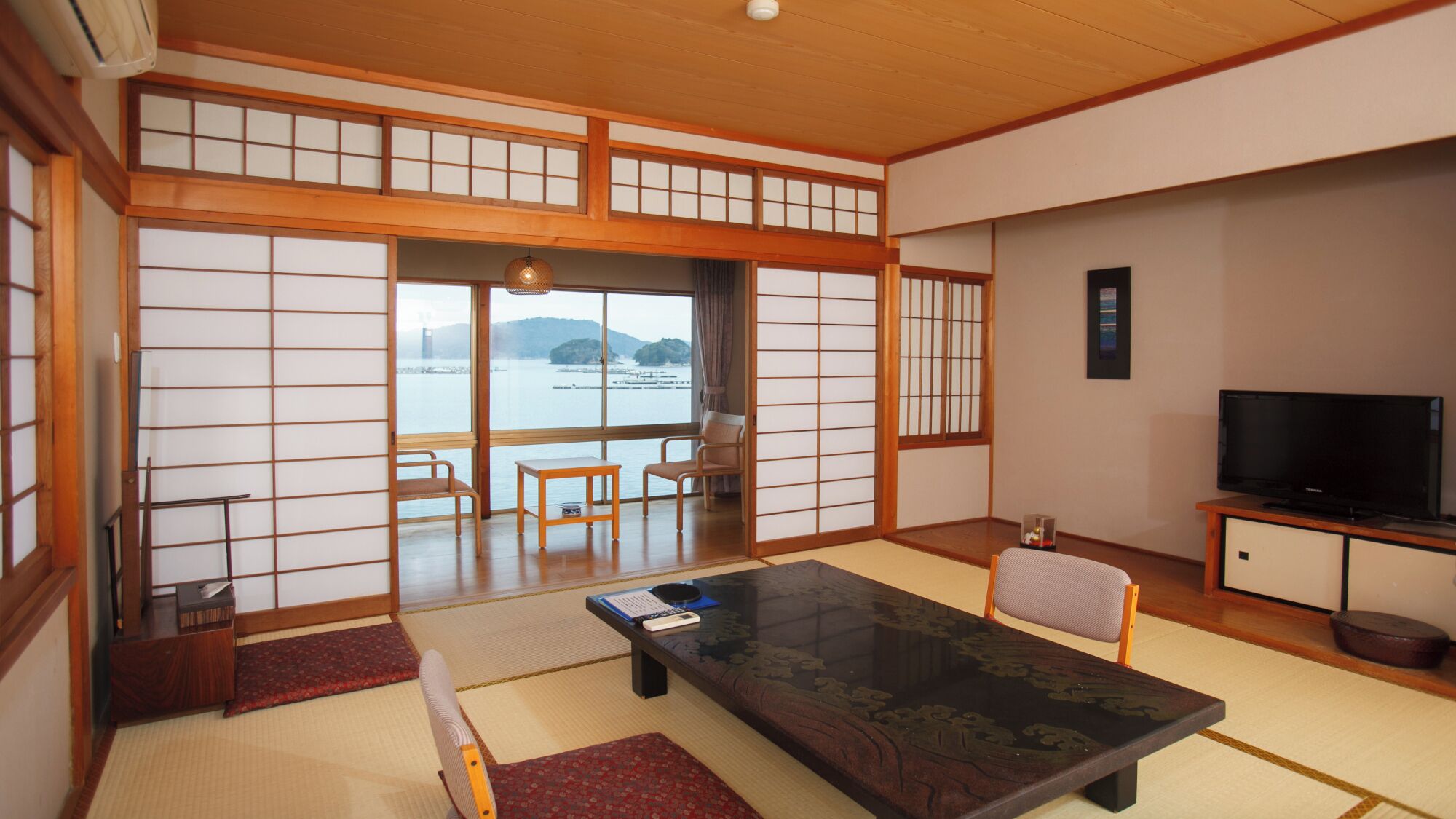 A traditional Japanese-style room with tatami flooring, a low table with cushions, a television, and a view of a body of water.
