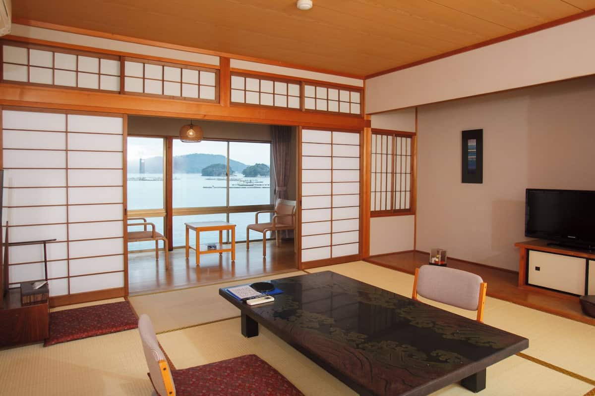 A traditional Japanese-style room with tatami flooring, a low table with cushions, a television, and a view of a body of water.