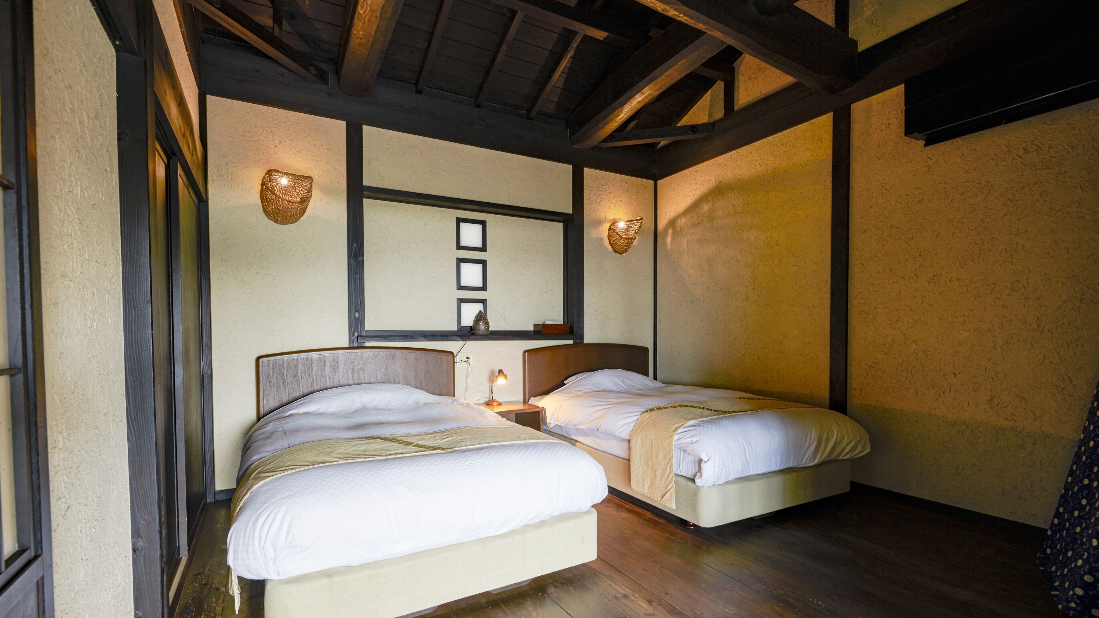 Een kamer met twee bedden, houten balken in het plafond, een wandplank met decoratieve objecten, en een tafeltje met een lamp.