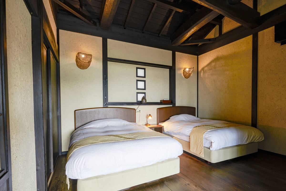 Une chambre avec deux lits, des poutres en bois au plafond, une étagère fixée au mur avec des objets décoratifs, et une petite table avec une lampe.