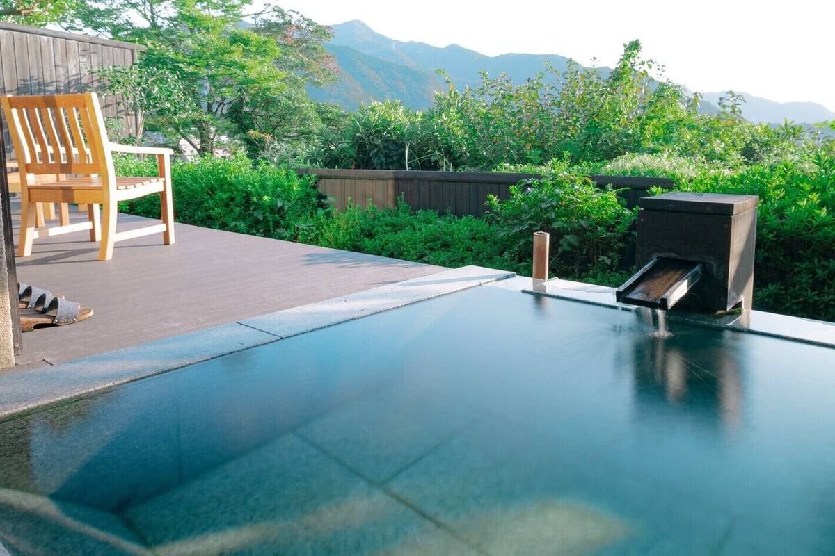 Une piscine avec une chaise en bois et une structure en bois, entourée de verdure et avec des montagnes en arrière-plan.