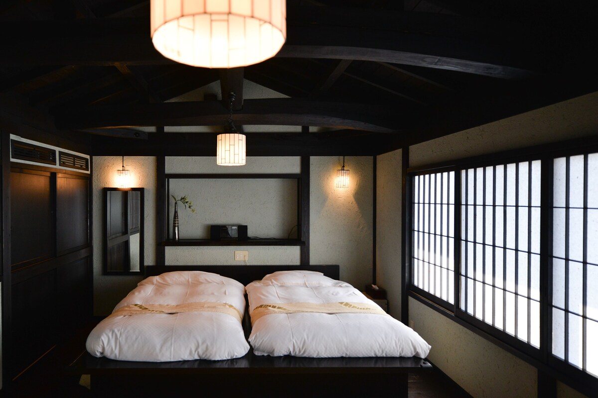 Habitació d'estil japonès tradicional amb sòl de tatami, un llit baix amb roba de llit blanca, una làmpada penjant i una finestra amb persianes verticals.