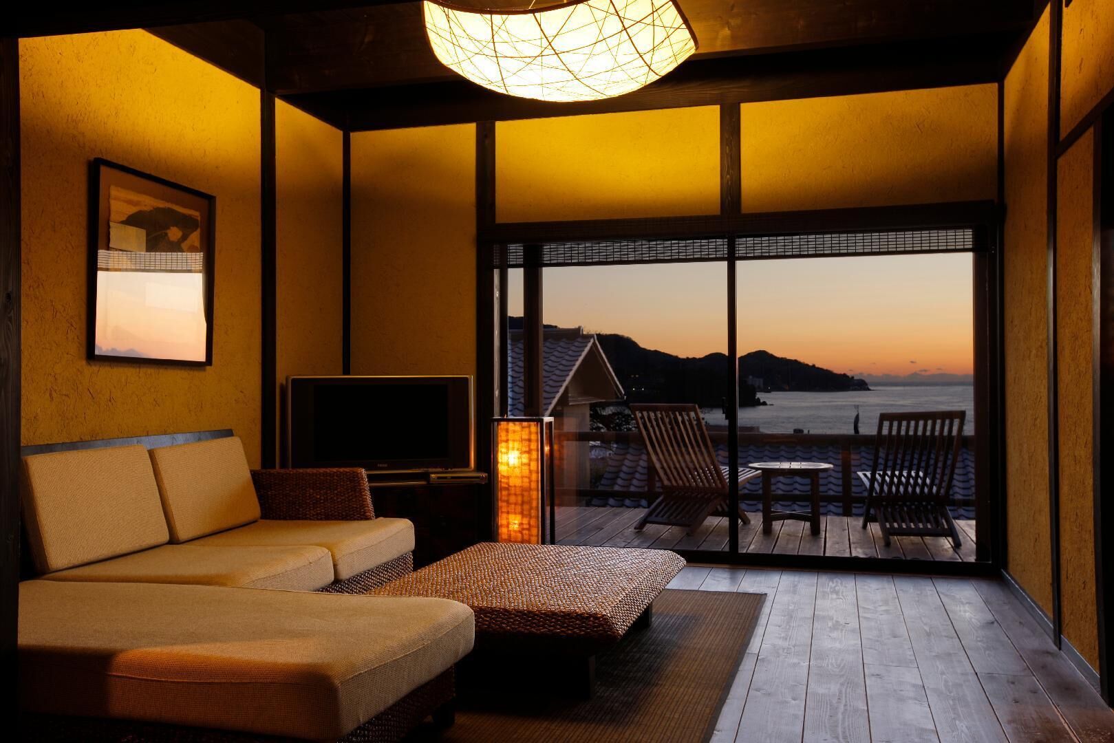Quarto com sofá, televisão, luminária e vista para o mar ao pôr do sol.