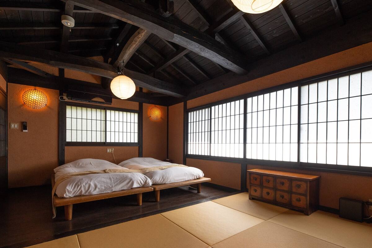 Une pièce de style japonais traditionnel, avec un sol recouvert de tatamis, un lit en bois, une commode et de grandes fenêtres à carreaux.