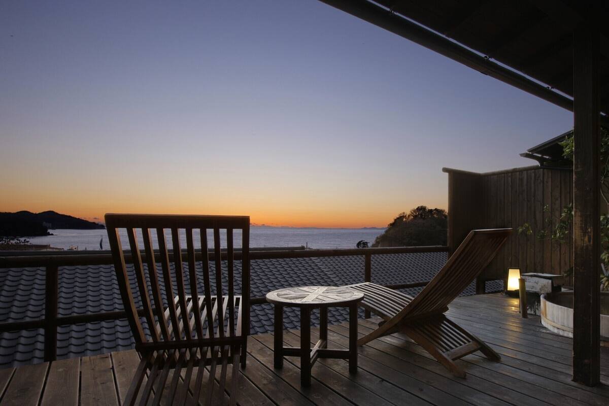 Une terrasse en bois avec une table et des chaises, donnant sur une étendue d’eau au coucher du soleil.