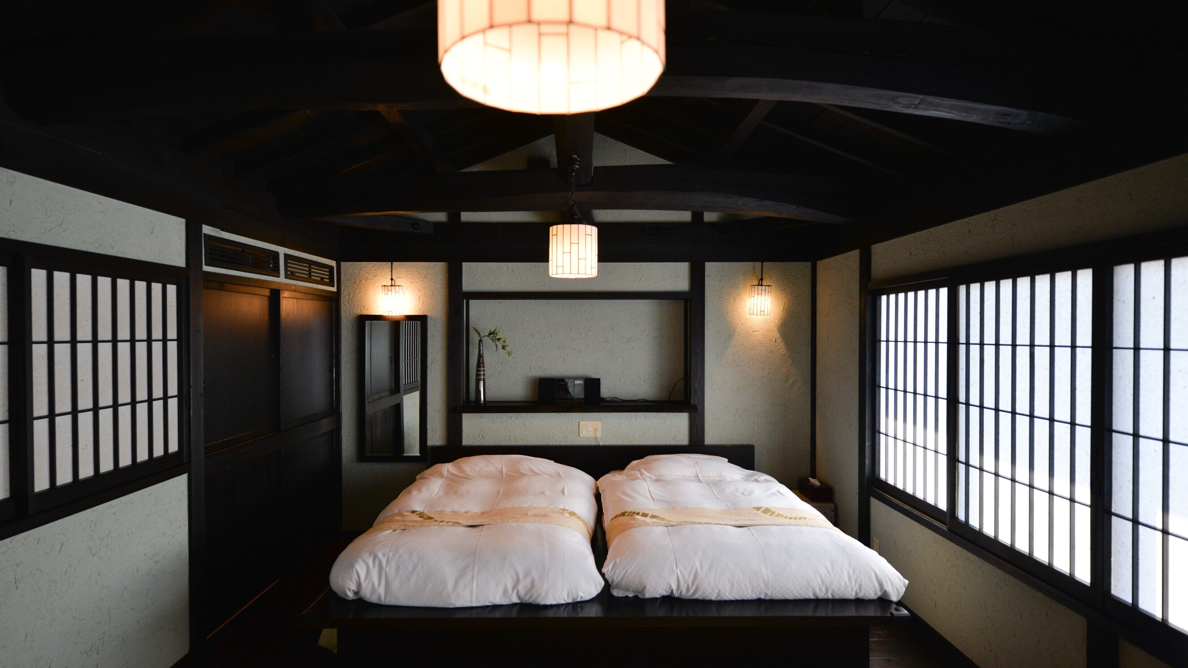 Een traditionele Japanse kamer met tatami vloeren, een laag bed met witte beddengoed, een hanglamp en een raam met verticale jaloezieën.