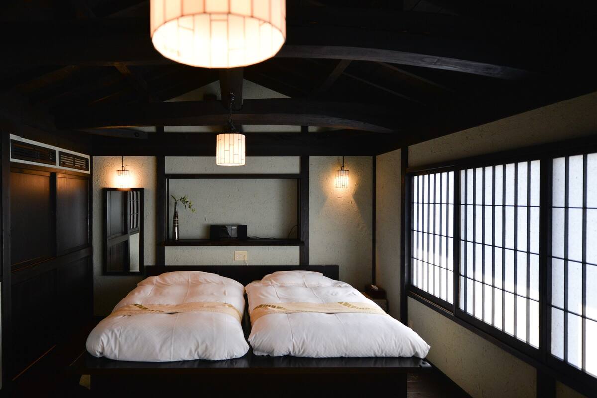 Une pièce de style japonais traditionnel, avec un sol recouvert de tatamis, un lit bas avec une literie blanche, une lampe suspendue et une fenêtre équipée de stores verticaux.