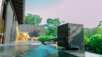 Una fuente de agua con un caño metálico, una silla de madera y un edificio al fondo.