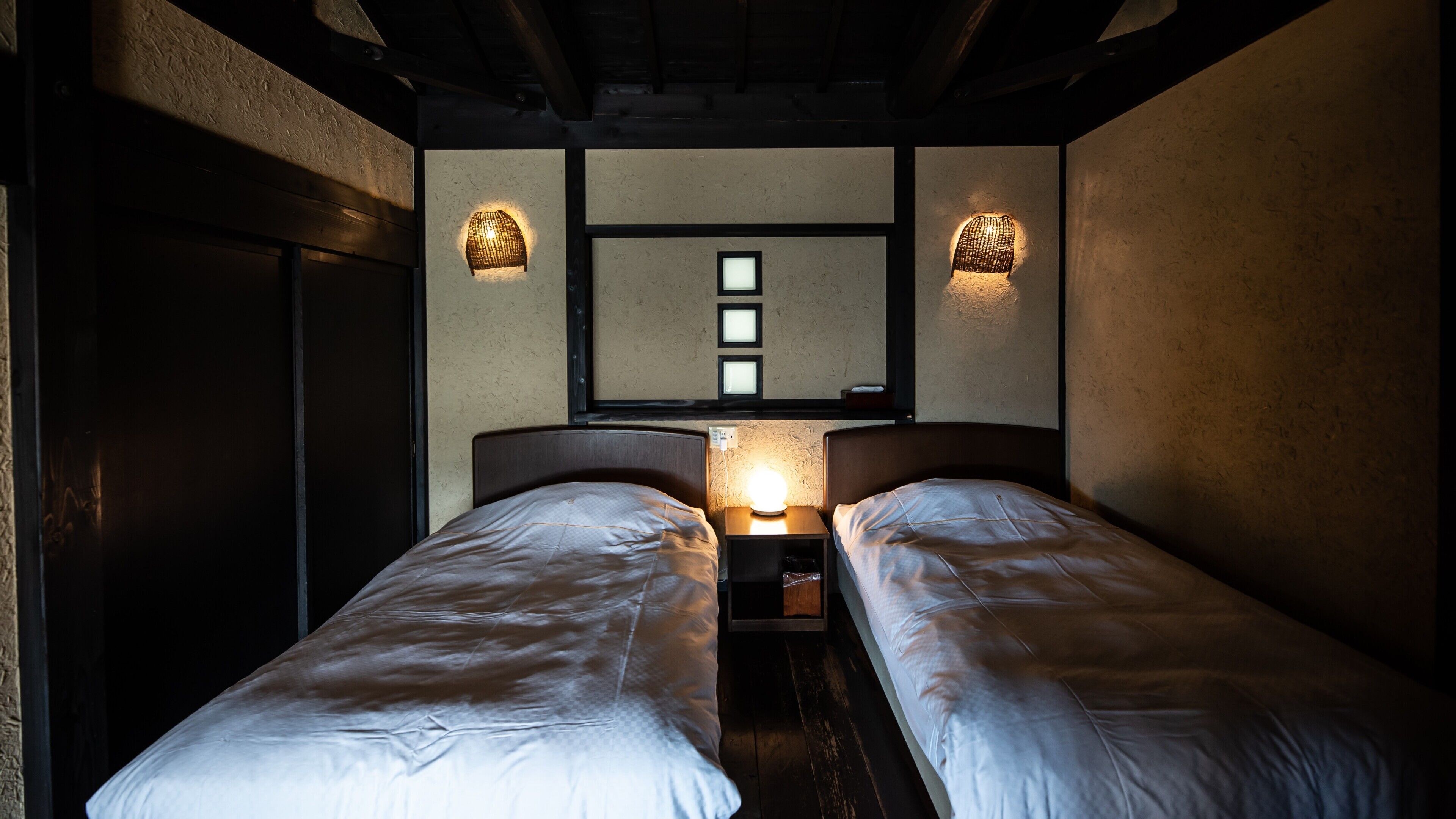 Een kamer met twee bedden, een wandlamp en een raam met jaloezieën.