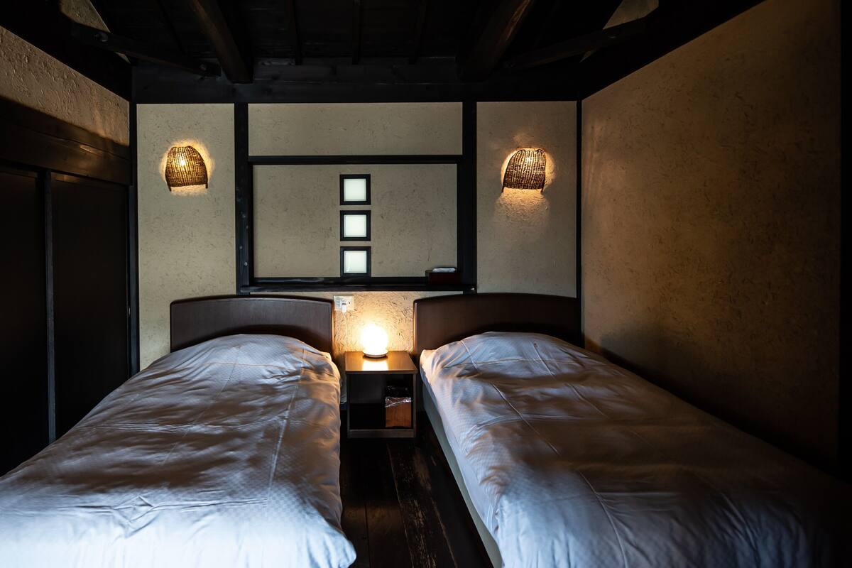Une chambre avec deux lits, un luminaire fixé au mur et une fenêtre avec des stores.