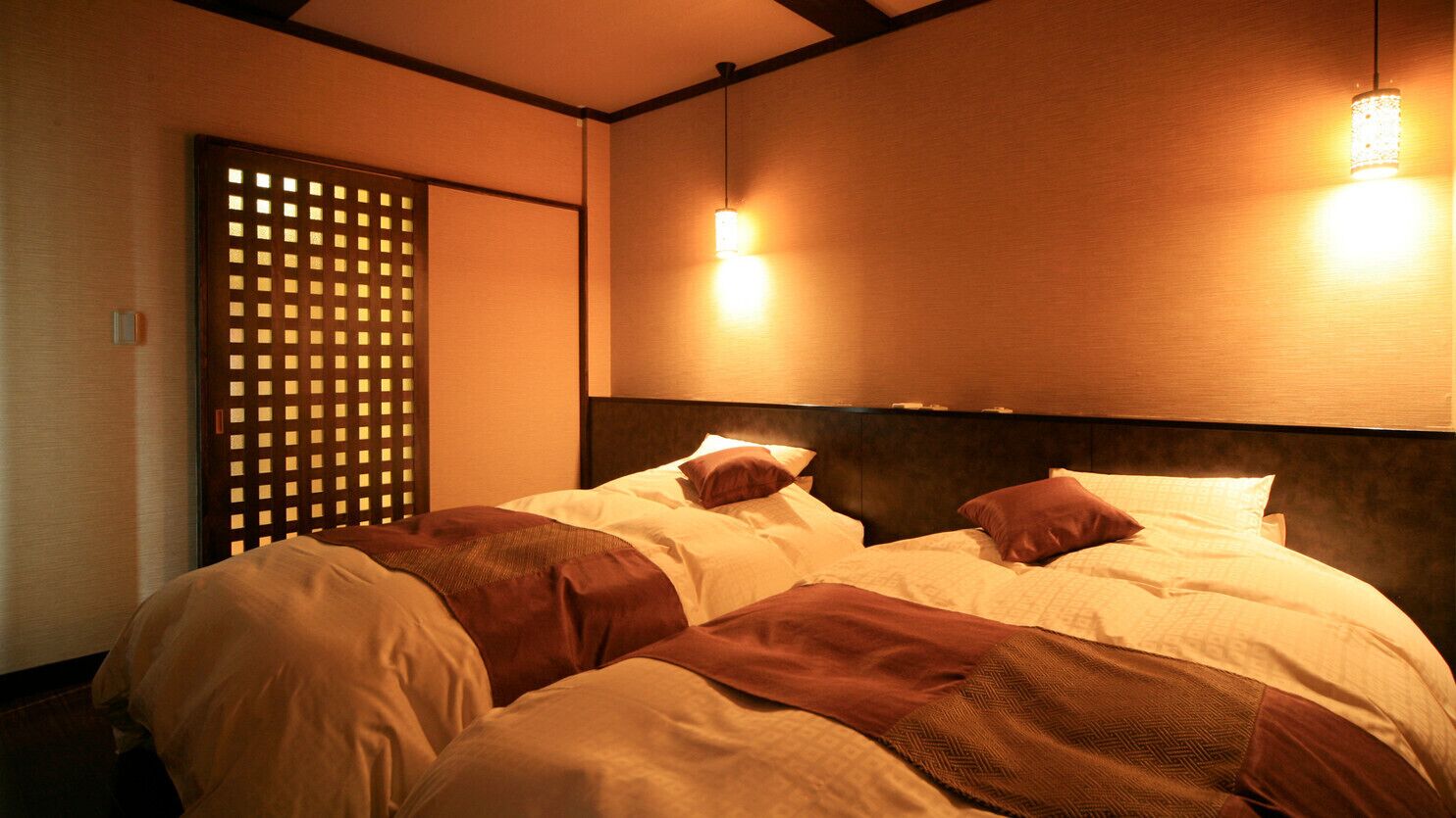 Quarto de hotel com duas camas, cabeceira de madeira e luminária fixada na parede.