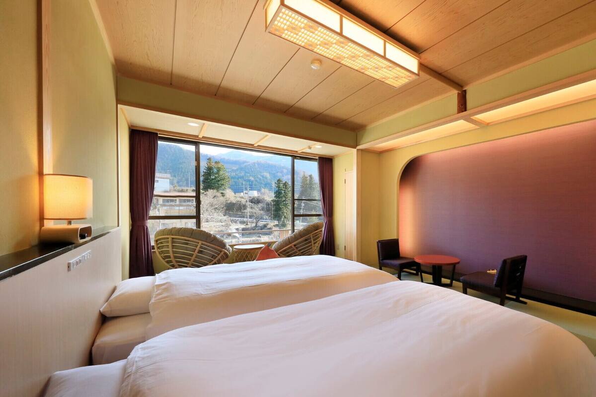 A hotel room with a large bed, a small table, two chairs, a view of the city, and a large window.