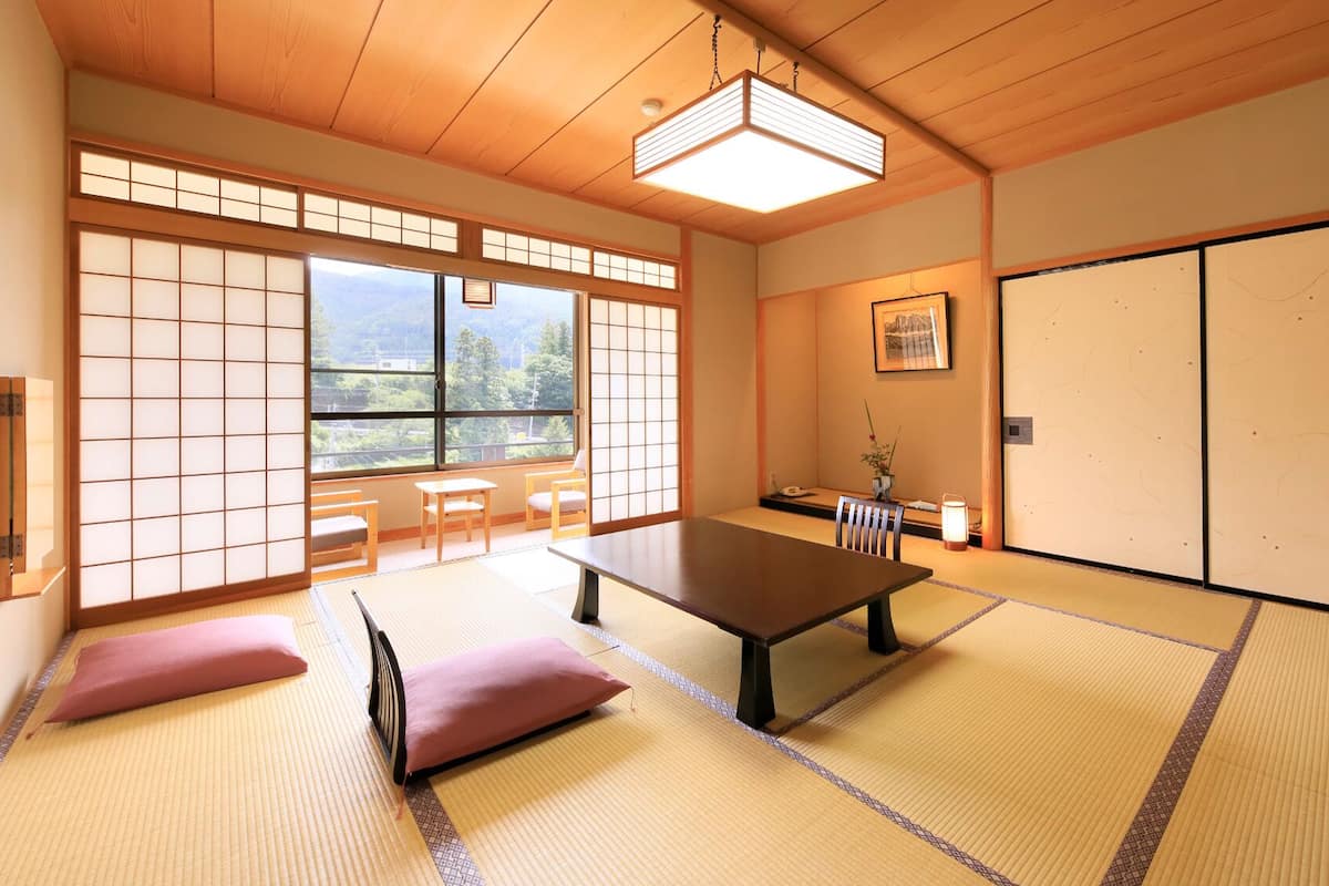 Une pièce de style japonais traditionnel, avec un sol recouvert de tatamis, une table basse, des coussins, des portes coulissantes et une vue sur la verdure.