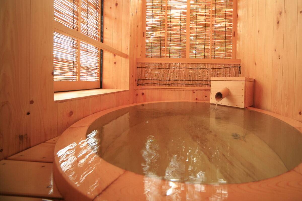 A wooden hot spring bath with a wooden interior, a window with bamboo blinds, and a small wooden structure with a hole.