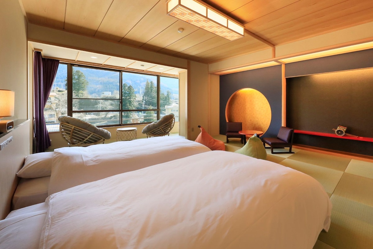 A hotel room with two beds, a large window, a round table, and a wall-mounted TV.