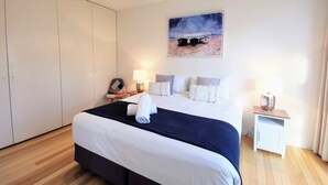 Una cama bien hecha con ropa de cama blanca y azul marino, un cabecero y una toalla enrollada sobre la cama. Se aprecia una mesita de noche con una lámpara, un pequeño tocador con lámpara y un cuadro en la pared que representa una playa.