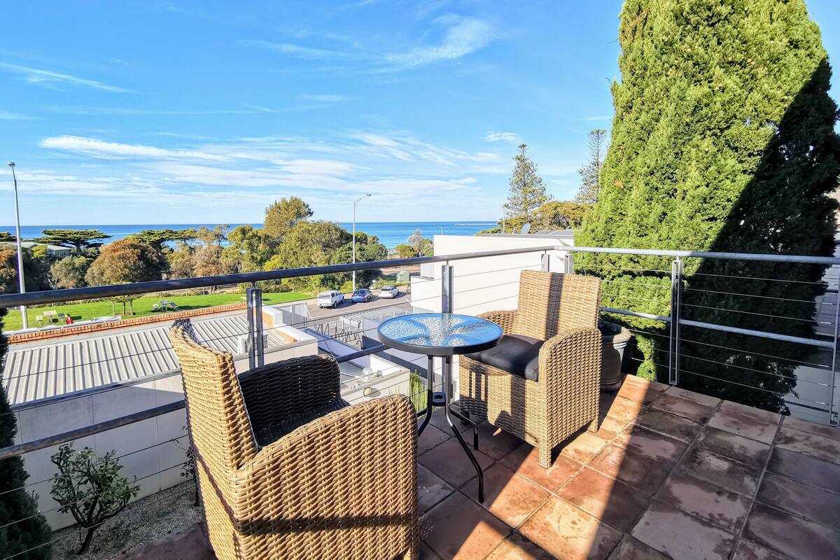 A balcony with wicker furniture, a round table, and a view of the sea and greenery.