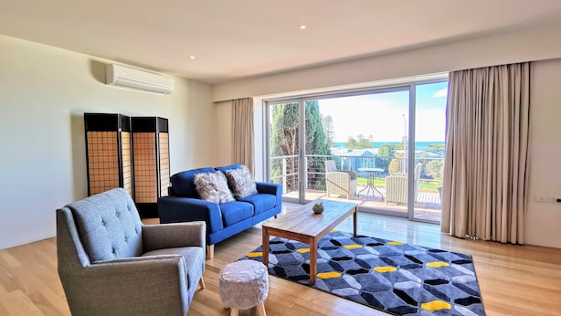Una sala moderna con un sofá azul, un sillón gris y una mesa de centro de madera. Una alfombra estampada cubre el suelo, y hay un ventanal corredero que da a un balcón con mobiliario exterior.