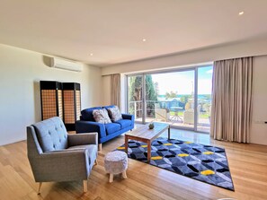 A modern living room with a blue sofa, grey armchair, and a wooden coffee table. A patterned rug is on the floor, and there is a sliding glass door leading to a balcony with outdoor furniture. - Phoenix Apartments (Lorne)