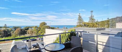 En balkon med fletmøbler, et rundt bord og udsigt til havet og grønne områder.