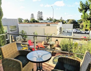 A balcony with wicker furniture, a glass table, and a view of a parking area and buildings. - Phoenix Apartments (Lorne)