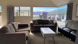 Un salon moderne avec un canapé, un fauteuil et une table basse. Une grande fenêtre offre une vue sur les montagnes et un immeuble.